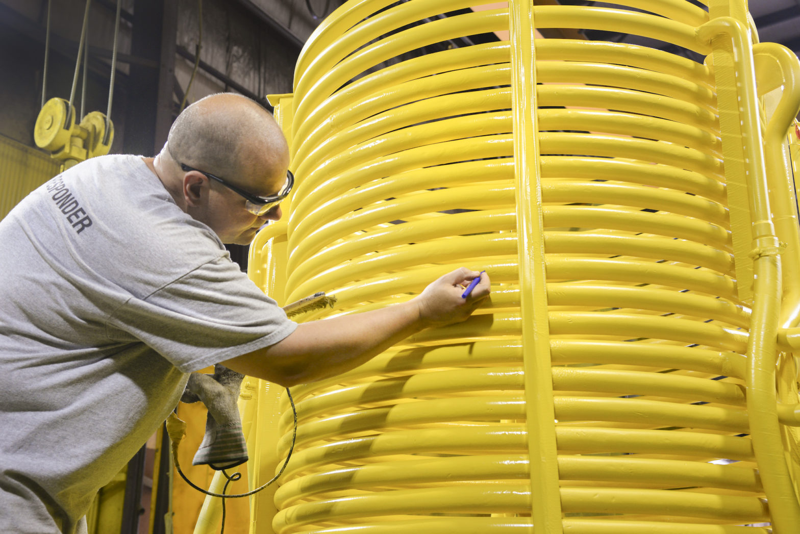 Trabalhador inspeciona visualmente um grande equipamento industrial com tubulações amarelas, utilizando uma ferramenta manual para verificação detalhada.